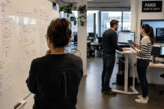 Mid-career female IT engineer stands thoughtfully at a whiteboard filled with technical notes in a softly lit French tech office, with two colleagues nearby in muted discussion and computer screens glowing in the background.