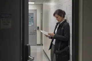 Female engineer in a lab hallway, reviewing notes alone, shot candidly through a partially open door with soft overhead lighting.
