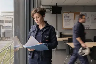 Female engineer reviewing notes by a window in an industrial break area, natural light, hint of workplace bustle in background.
