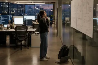 Young female engineer in open-space office studying technical plans on glass wall at dusk, ambience of focus and late work