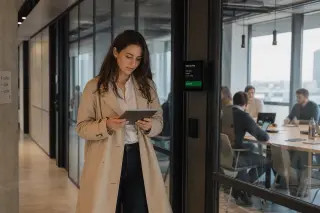 Woman engineer standing in a corporate hallway, thoughtfully reading job offers on a tablet, modern office setting with glass and natural light.